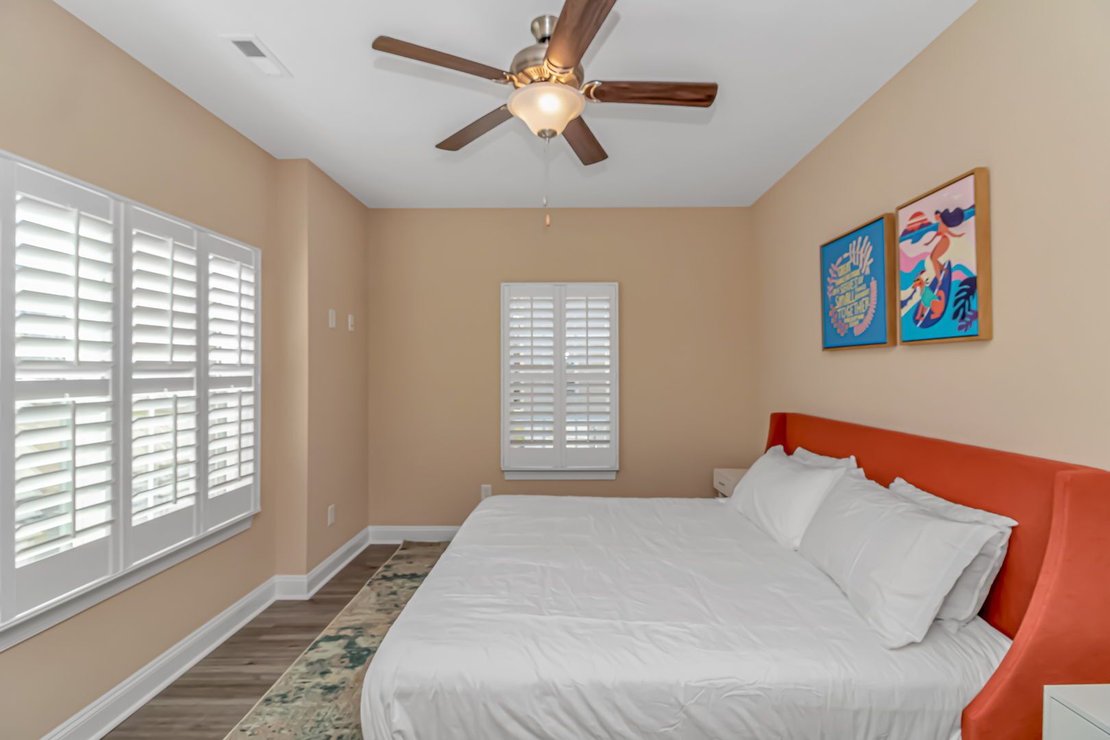 A bedroom with a bed, a ceiling fan, two windows with blinds, and two wall-mounted paintings.