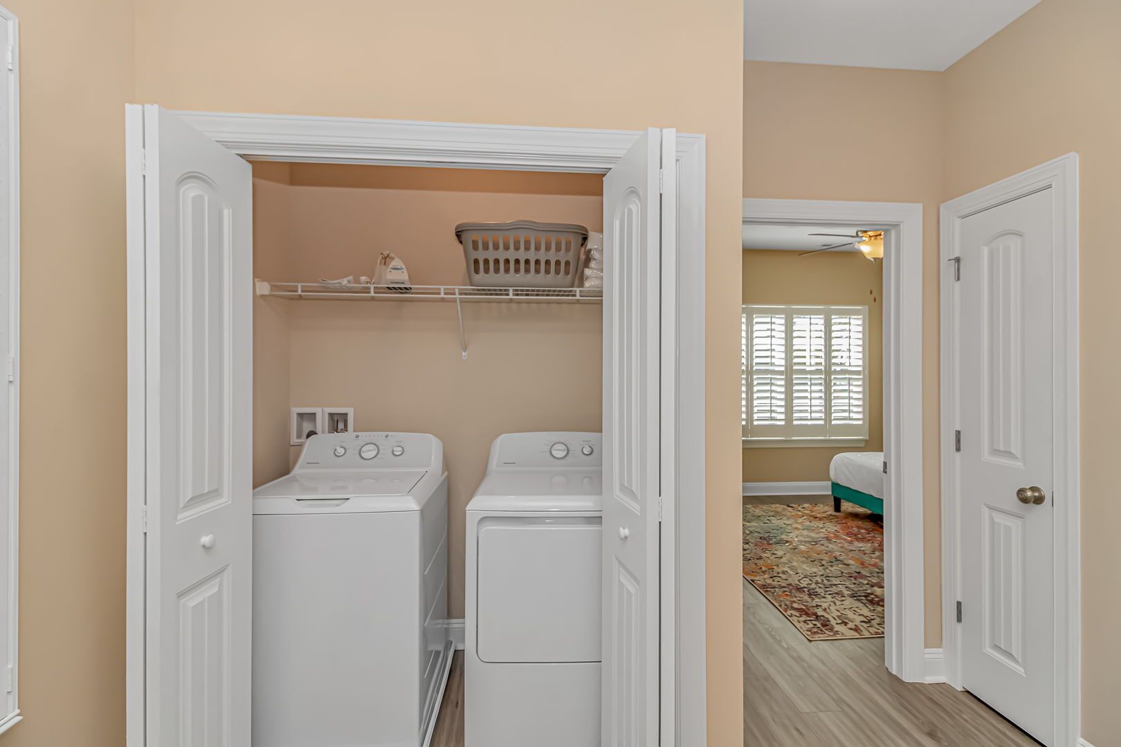 Une buanderie avec deux machines à laver et deux sèche-linge blancs, un panier posé sur une étagère, et une porte donnant sur une chambre à coucher avec un lit et une fenêtre.