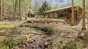 Ein rustikales Blockhaus mit einer Holzterrasse und einem kleinen Bach in bewaldeter Landschaft.
