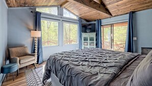 Ein Schlafzimmer mit einem großen Bett, einem Sessel, einem Fernseher und Blick ins Freie durch große Fenster.