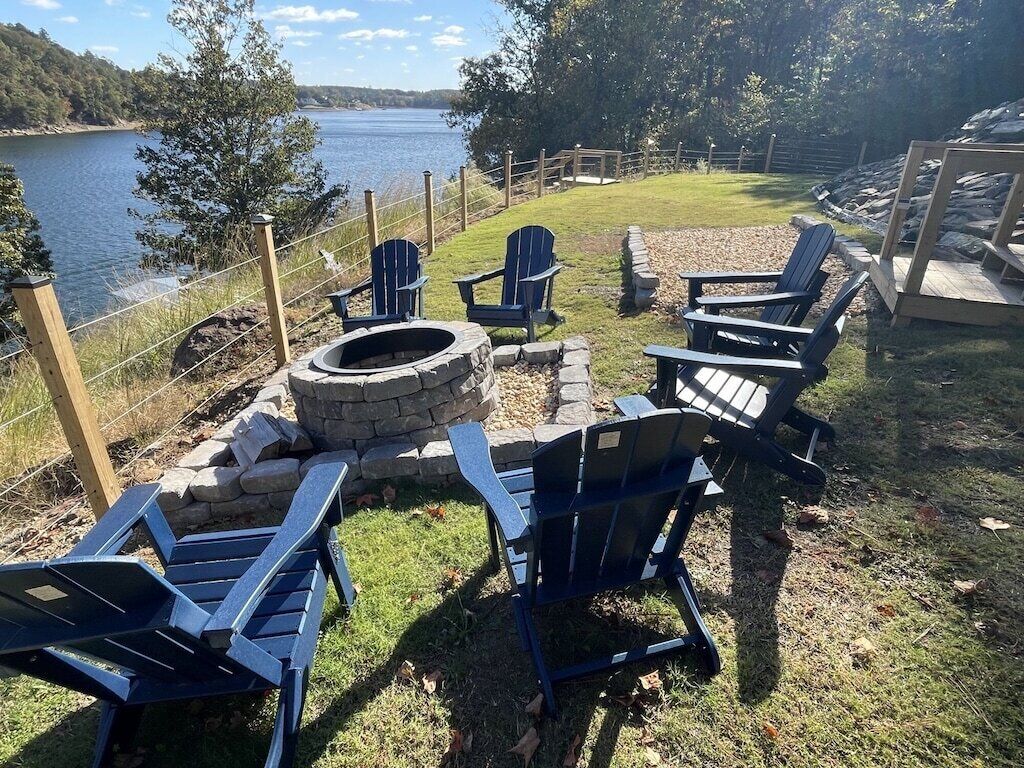 Un área de estar al aire libre frente al lago, con sillas tipo Adirondack y un hogar a leña de piedra.