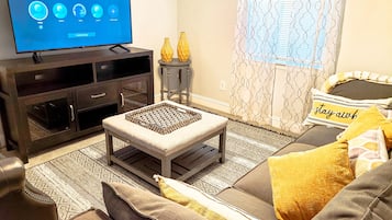 Een woonkamer met een flatscreen-tv waarop een profielselectiescherm wordt weergegeven, een donker houten tv-meubel, een kamerkleed met dessin en een bank met gele kussens.
