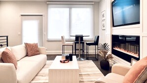 Een moderne woonkamer met een bank, salontafel en open haard. Er is een televisie aan de muur bevestigd en een raam met zonwering.