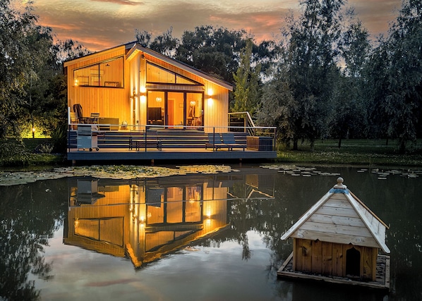 A wooden cabin with a deck, a dock, and a reflection in the water.