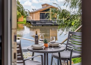 A dining table with a bottle and glasses, a chair, and a view of a house by a body of water.