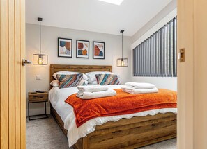 A bedroom with a wooden bed, a nightstand, a bedside table, a window with blinds, and three framed artworks on the wall.