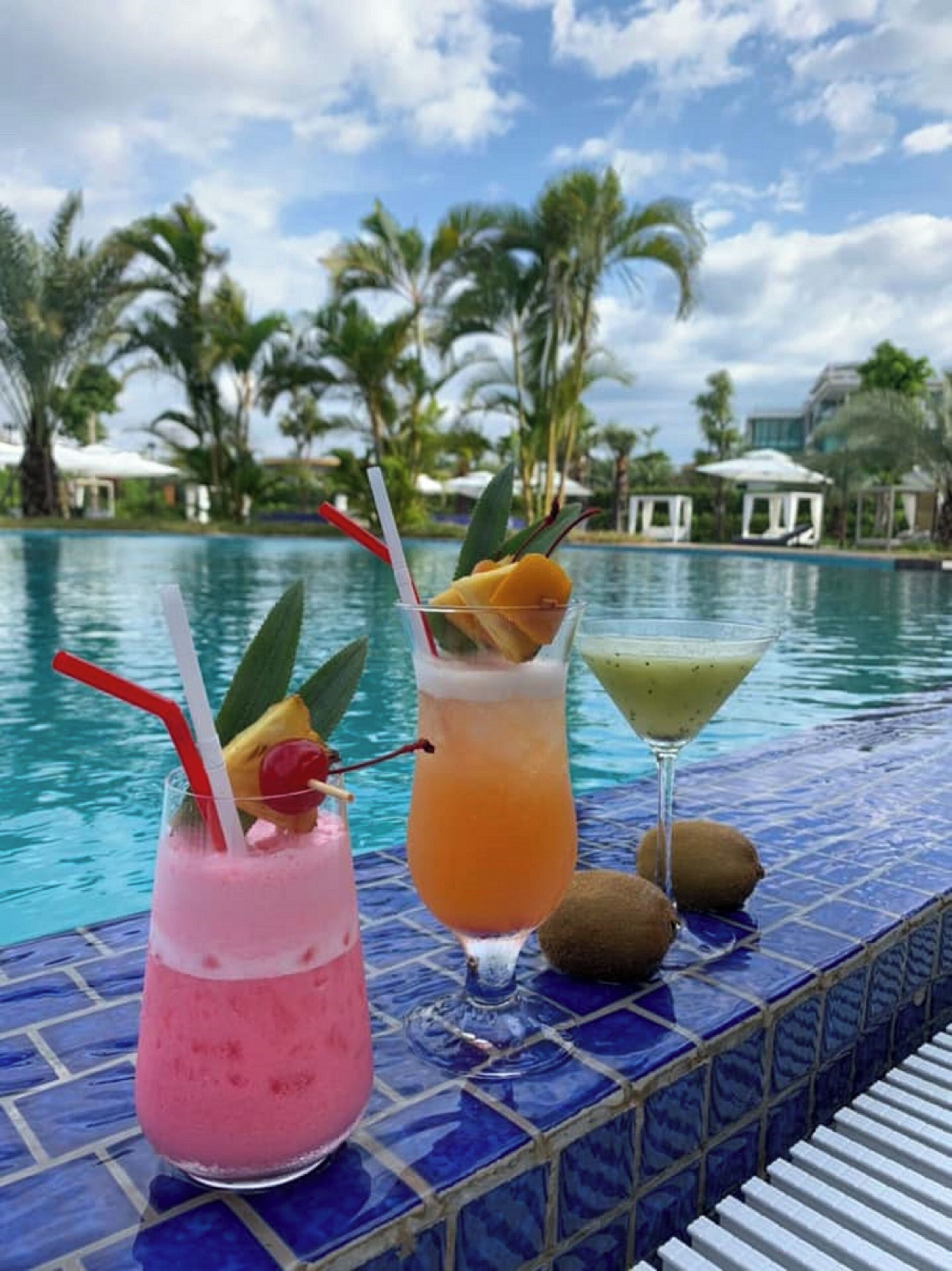 Drie cocktails op een tafeltje bij het zwembad, met een betegelde rand, palmbomen en een heldere lucht op de achtergrond.