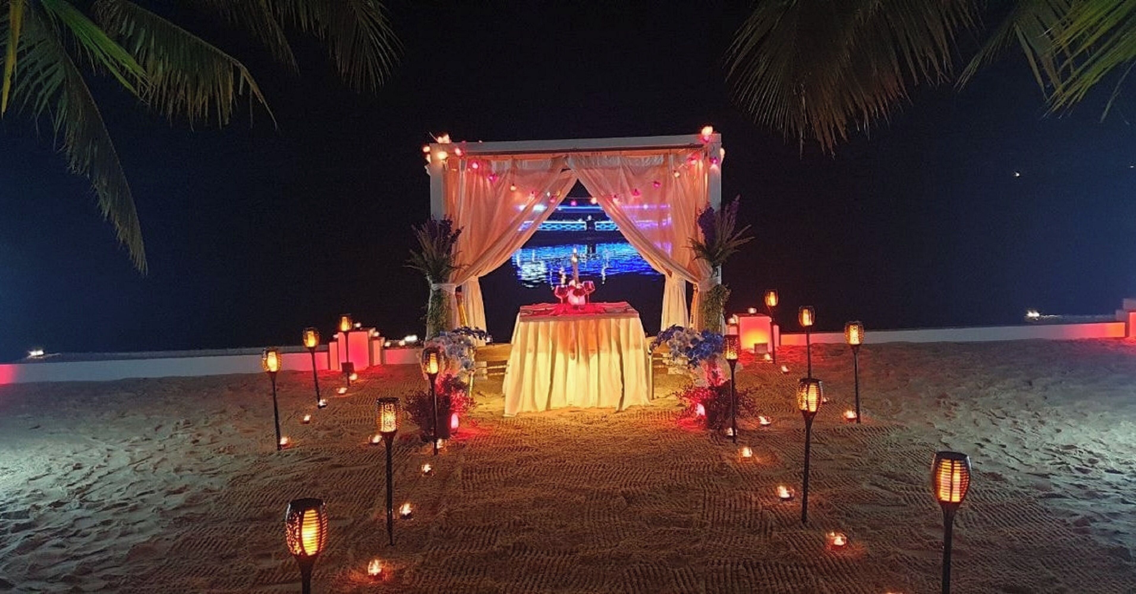 An outdoor wedding setup with a decorated arch, a table, and candles on a sandy beach at night.