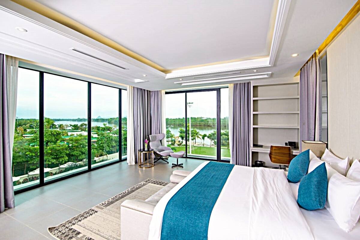 Un espacioso dormitorio con una cama grande, un escritorio y una silla, que ofrece vistas a un paisaje exuberante a través de amplios ventanales.