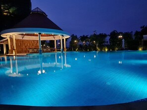 A large, well-lit swimming pool with a covered seating area and a building in the background.