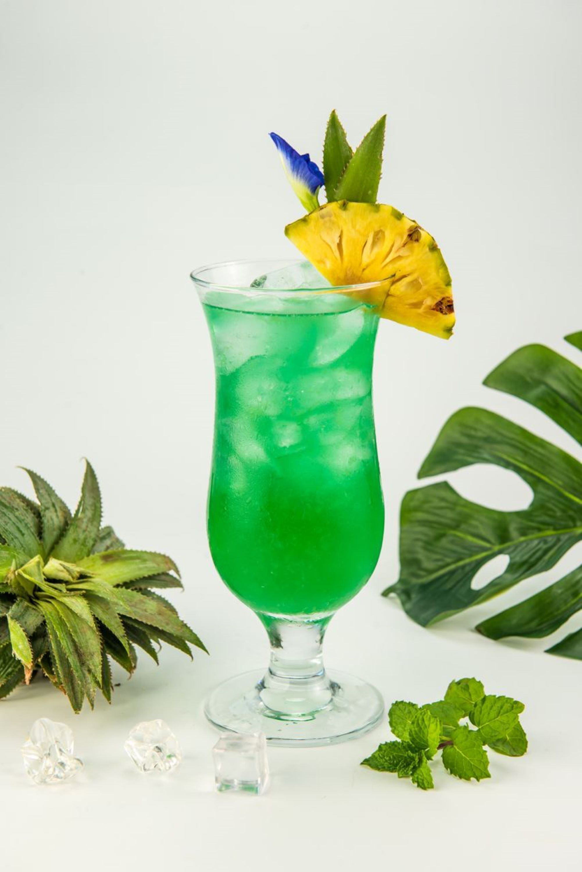 A green cocktail in a glass with a pineapple garnish, surrounded by green leaves and mint.