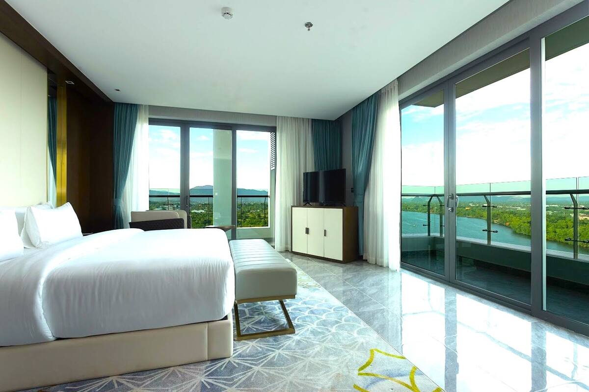 Una habitación de hotel moderna con una cama grande, zona de estar, televisión y balcón con vistas panorámicas.