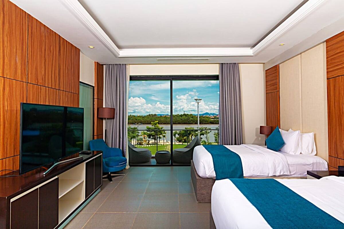 Habitación de hotel con televisor grande, dos camas, zona de estar con vistas al exterior y balcón con panorama.