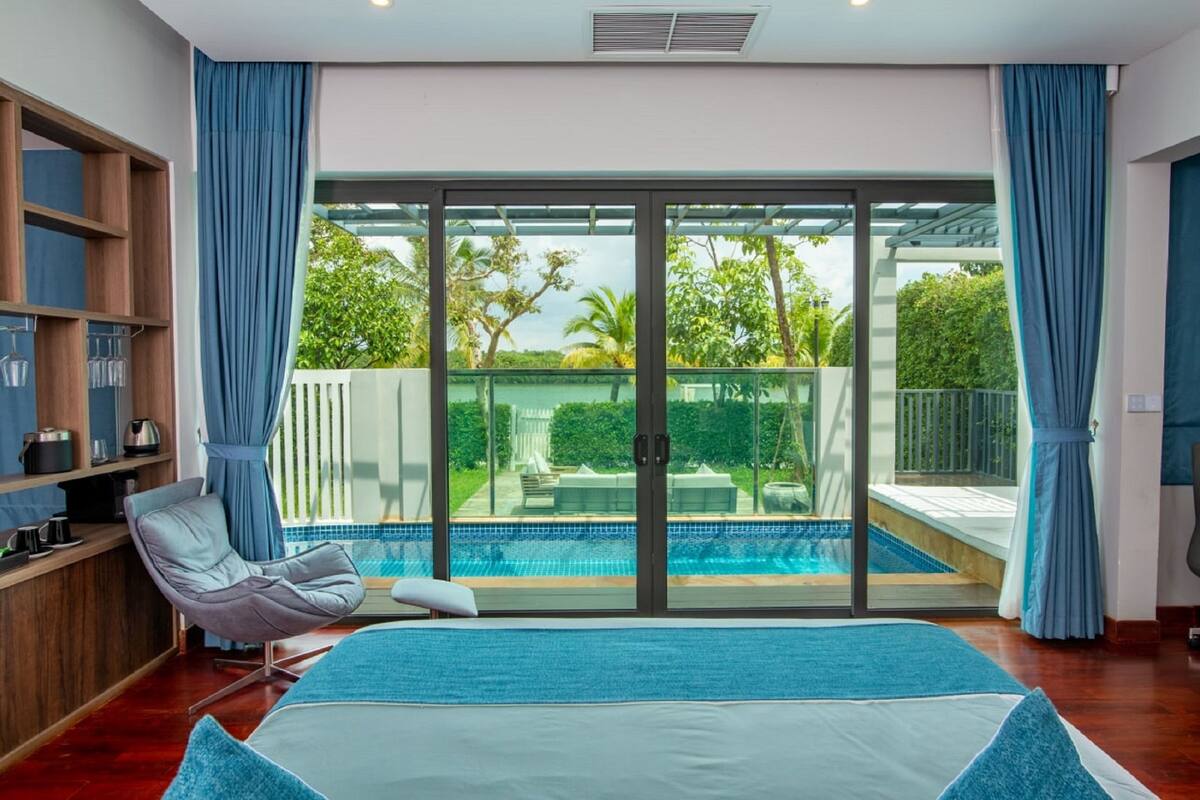 Un dormitorio con una cama grande, una silla, un estante de madera y vistas a una piscina y vegetación al exterior.
