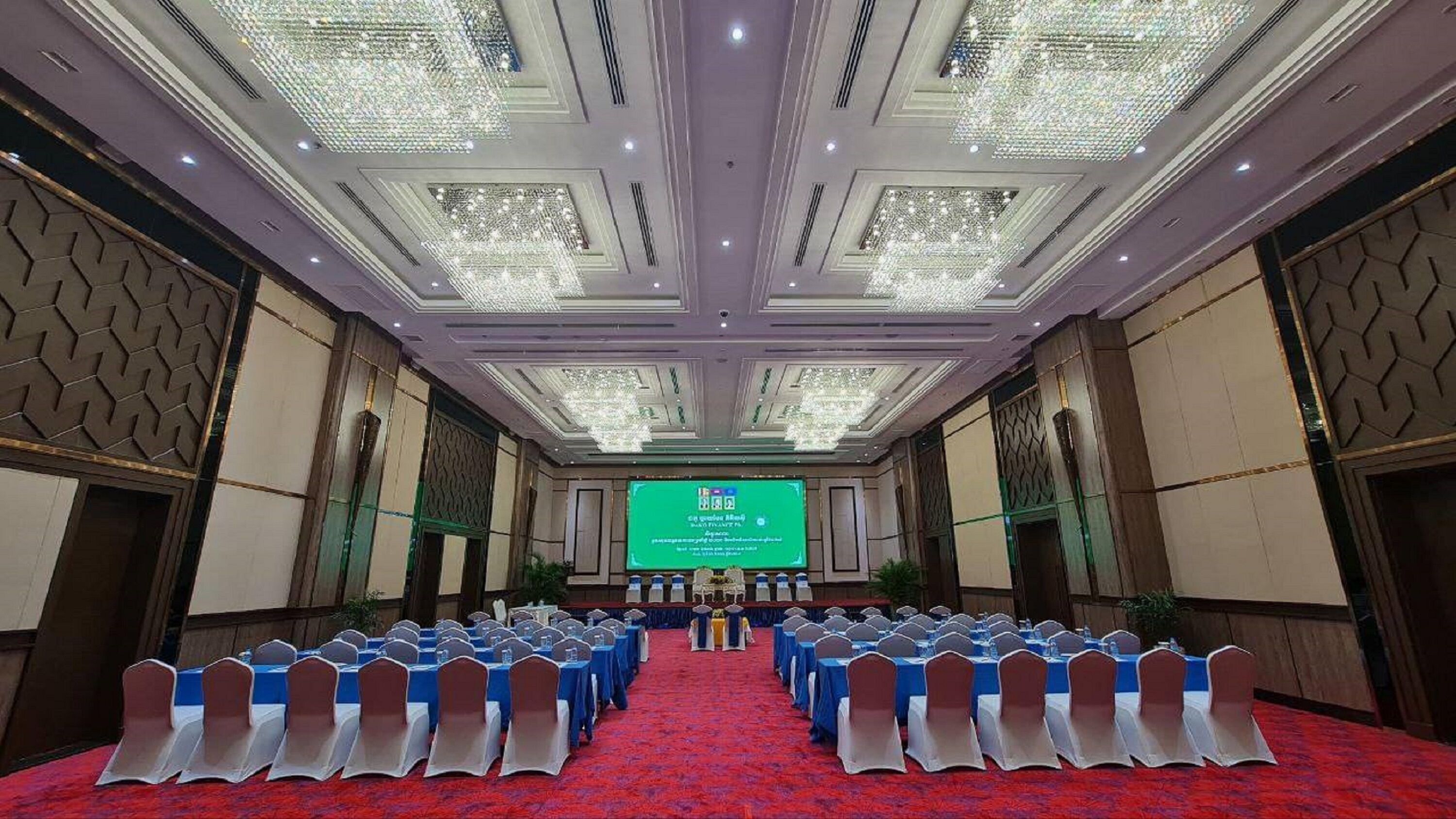 Un salón de conferencias con filas de mesas y sillas, una pantalla grande al frente y un techo alto con candelabros.