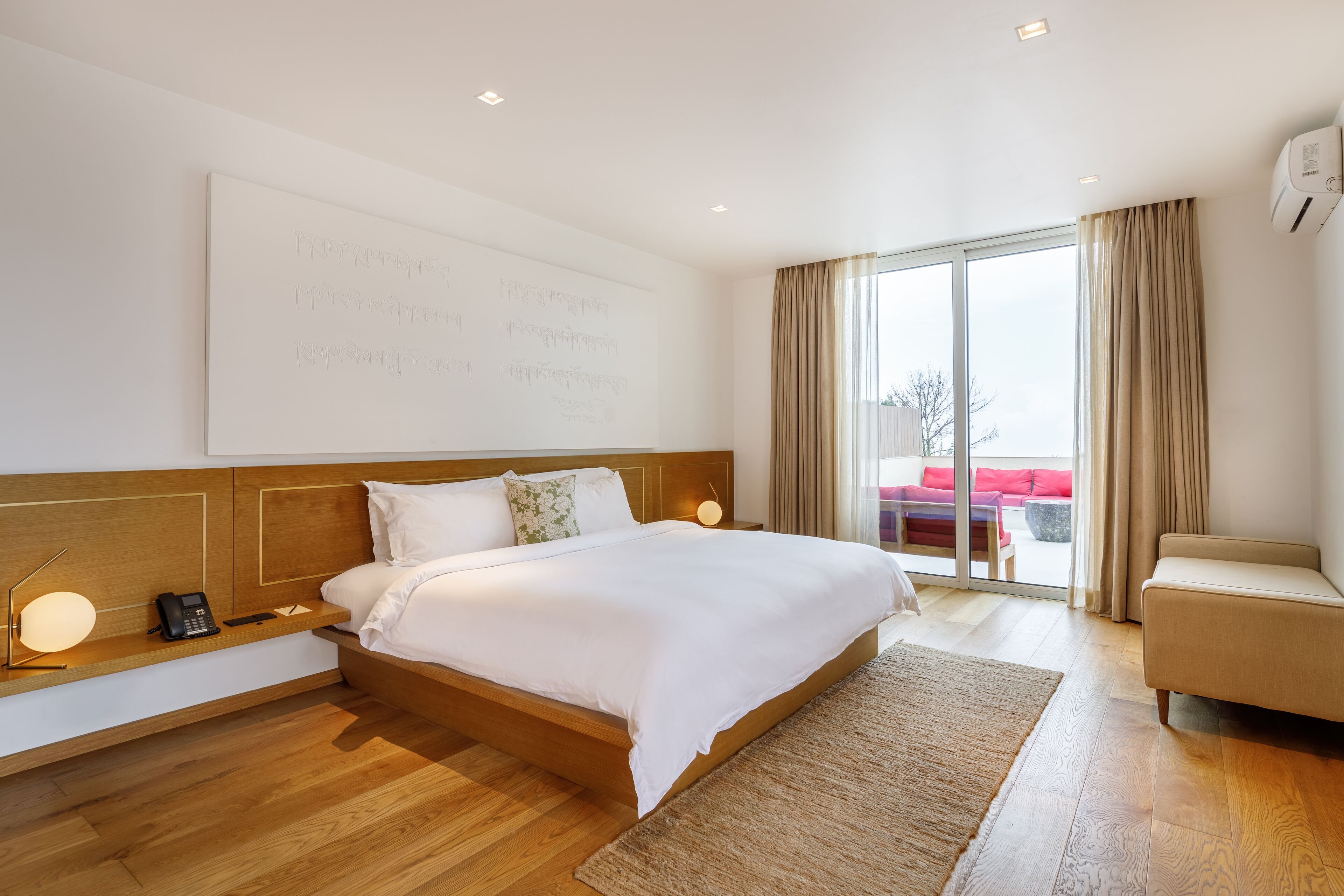 A modern hotel room with a large bed, wooden headboard, bedside table with a lamp, and a view of the outdoors through a sliding glass door.