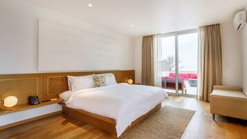 A modern hotel room with a large bed, wooden headboard, bedside table with a lamp, and a view of the outdoors through a sliding glass door.