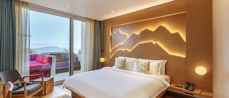 Un dormitorio moderno con una cama grande, cabecera de madera con diseño de montaña, una silla y vista a un sofá rojo a través de una puerta de vidrio.