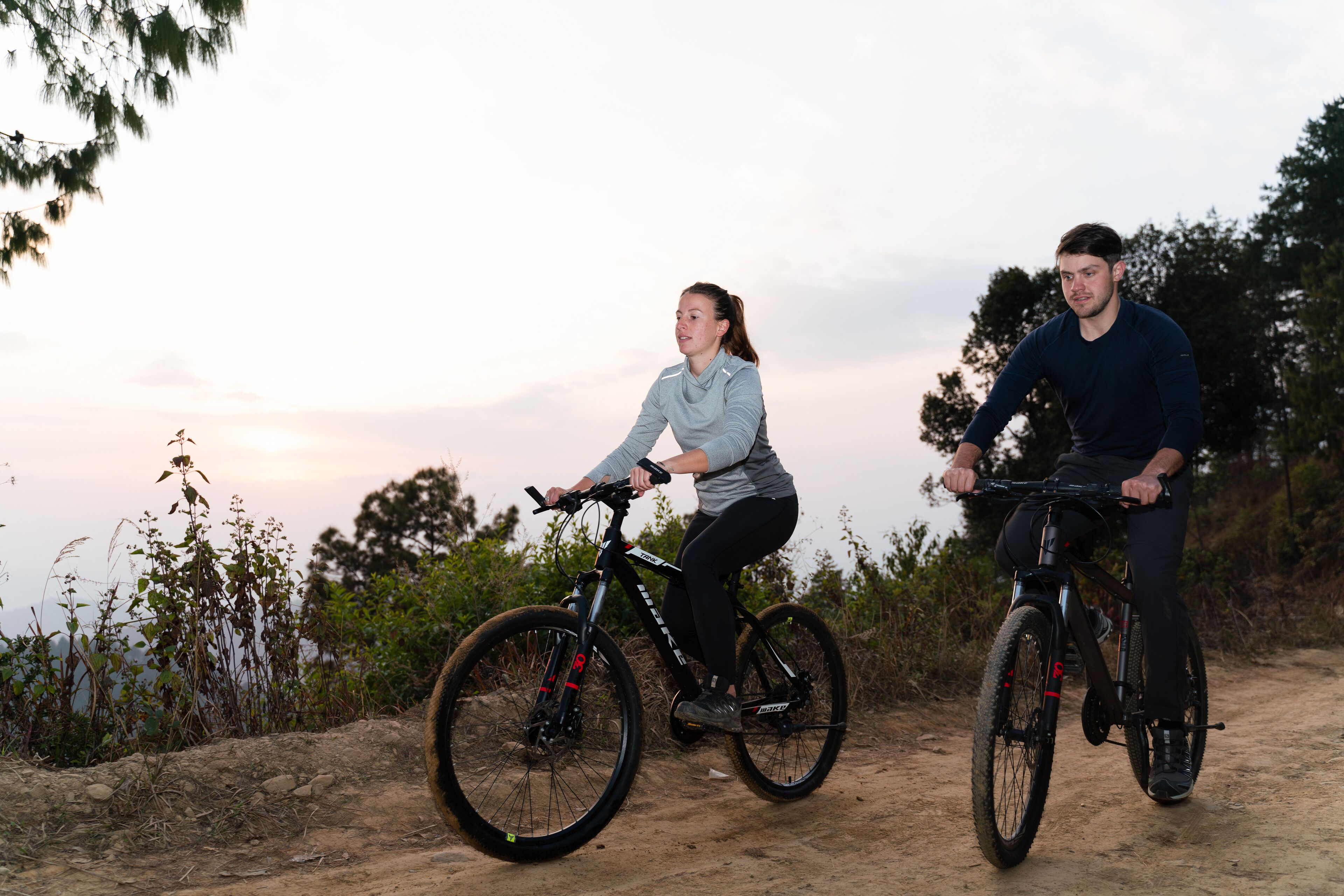 Two people riding bicycles on a dirt path with trees and a sunset in the background.
