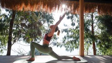 Una persona realizando una postura de yoga sobre una cubierta de madera, con vistas a árboles y un techo de paja.