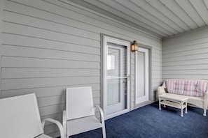 Eine überdachte Veranda mit blauem Teppich, weißen Stühlen, einem Korbsessel und einer Tür mit Leuchte.
