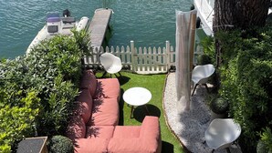 Un puerto deportivo con barcos amarrados, una terraza de madera con un sofá y sillas, y edificios a lo lejos.