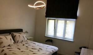A bedroom with a bed, a nightstand, a chair, a window with blinds, and a wall-mounted light fixture.