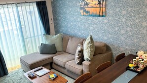 A modern living room with a sofa, coffee table, and dining table. There is a painting on the wall, a ceiling light, and an air conditioning unit. - PRIME TREE Izumizaki CONDO HOTEL (Naha)
