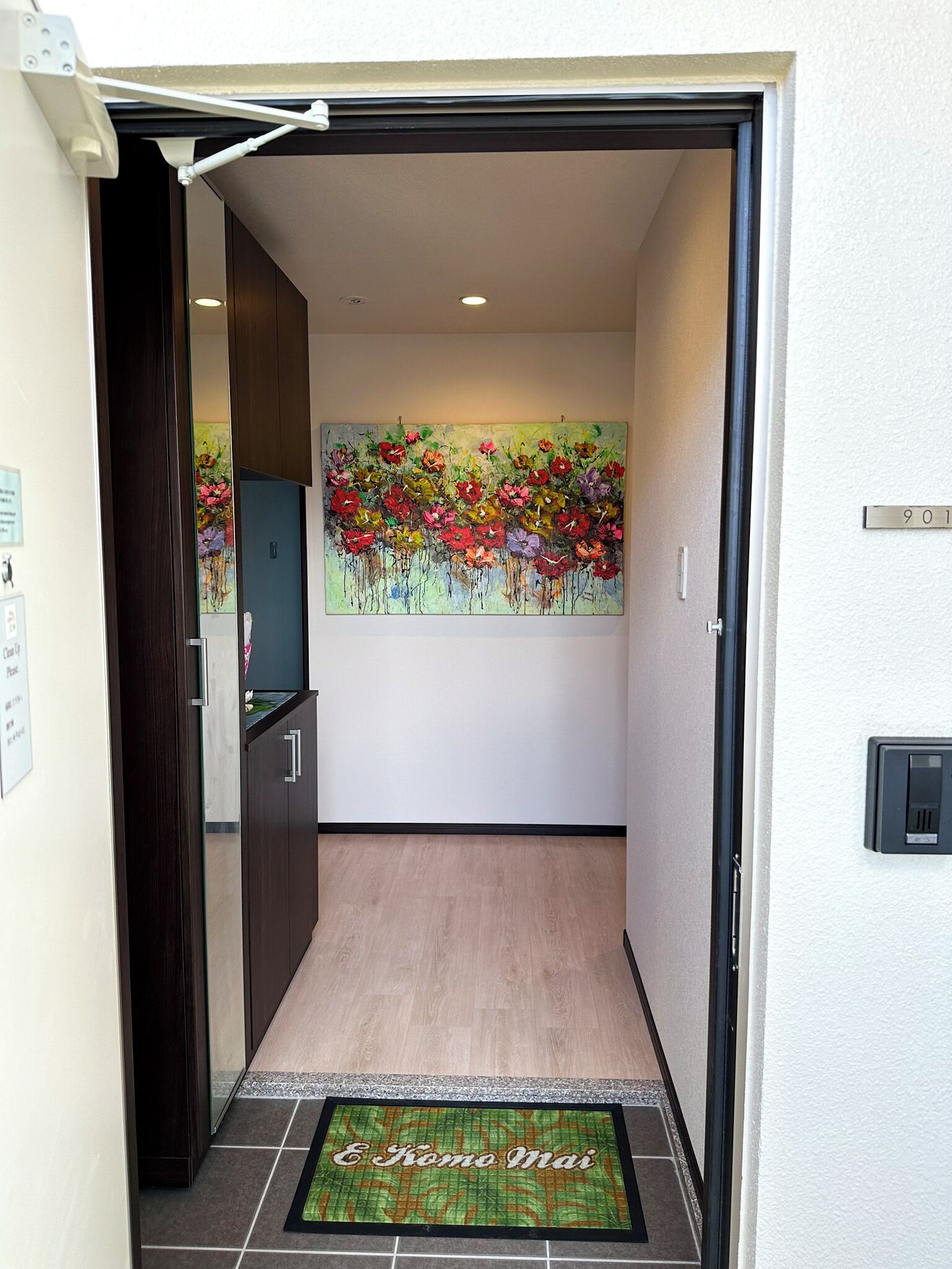 A hotel entrance with a wooden floor, a welcome mat, a painting of flowers, and a door with a number plate.