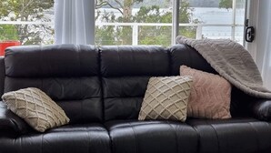 A black leather sofa with three textured throw pillows in a room with large windows offering a view of a lake and trees. - Peaceful -Local Wine & Tamar River Views/Pets Allowed (Clarence Point)