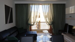 Ein Wohnzimmer mit einer schwarzen Couch, einem Couchtisch mit weißen Blumen, einem Fernseher und einem Balkon mit grünen Vorhängen.