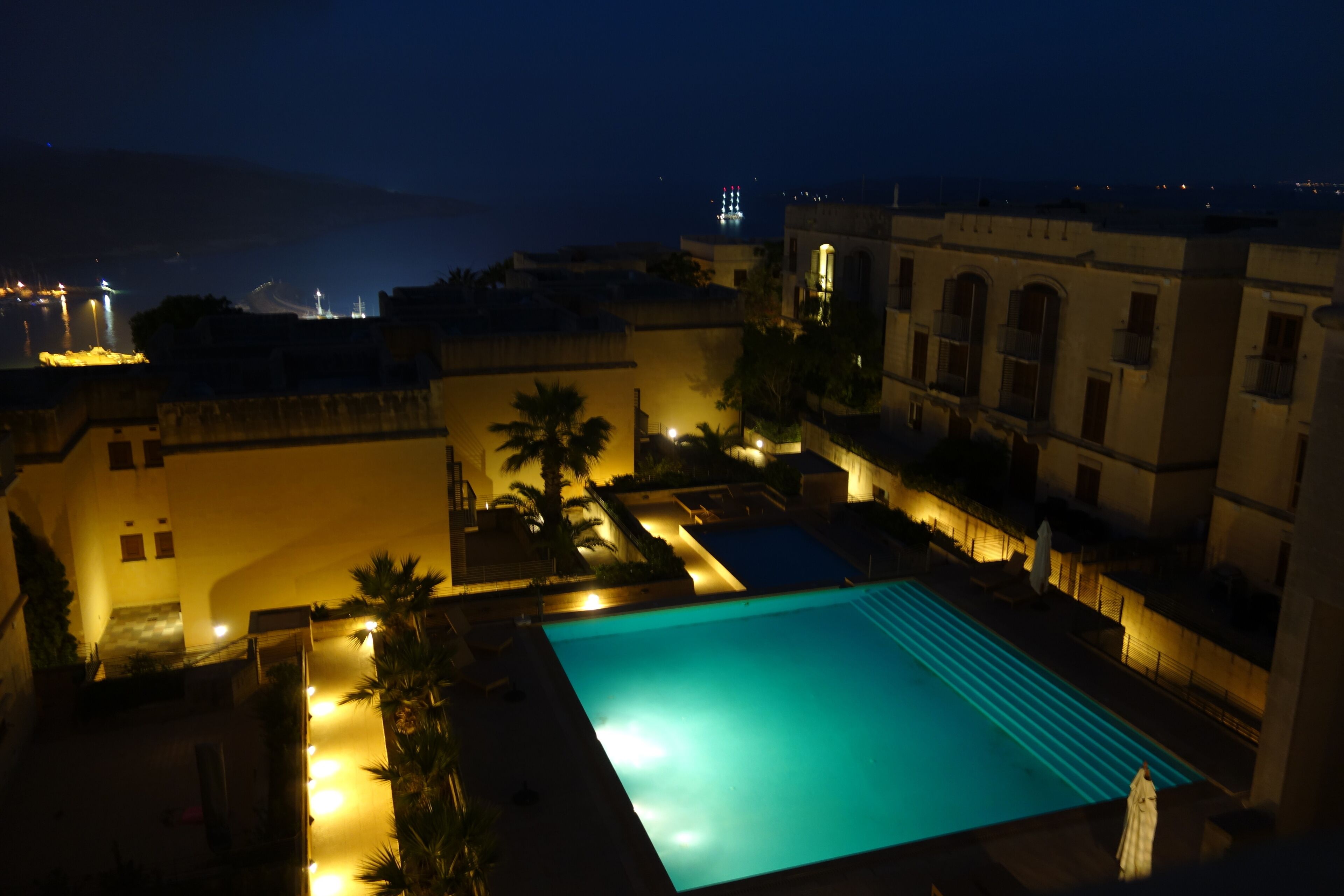 A nighttime view of a swimming pool area with surrounding buildings, including a pool with steps leading into it and a palm tree.