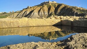 Eine Küstenlandschaft mit einem Hügel, auf dem sich eine schlossartige Struktur befindet, ruhigem Wasser, das die Szene spiegelt, und blauem Himmel.