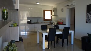 Eine moderne Küche mit weißen Schränken, eine Essecke mit weißem Tisch und schwarzen Stühlen sowie ein kleines Fenster mit Holzrahmen.