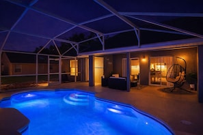 Een zwembad met een overdekte patio, een hangstoel en een goed verlichte binnenruimte.