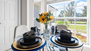 A dining table set for two with black plates and gold rims, a vase with sunflowers, and a view of a green landscape through a large window. - Beautiful 3BR Home Heated Pool | Ping-Pong |Disney (Davenport)