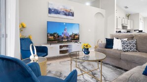 A modern living room with a flat-screen TV, blue and grey sofas, a glass coffee table, and a kitchen area in the background. - Beautiful 3BR Home Heated Pool | Ping-Pong |Disney (Davenport)