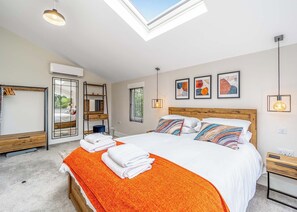 Ein Schlafzimmer mit Oberlicht, einem Bett mit bunten Kissen, einem Nachttisch, einem Spiegel und Kunstwerken an der Wand.