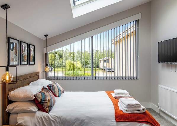 Ein modernes Hotelzimmer mit großem Fenster, einem Bett mit weißer Bettwäsche, einer roten Tagesdecke, einem wandmontierten Fernseher und gerahmten Bildern an der Wand.