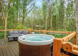 Ein Whirlpool auf einer Holzterrasse mit einem grauen Sofa und einem Tisch, umgeben von einem Wald.