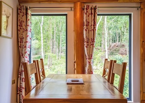 A dining table with chairs, large windows with curtains, and a forest view. - 2 bedroom accommodation in Ellesmere (Ellesmere)