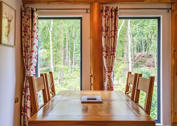 Mesa de jantar com cadeiras, janelas amplas com cortinas e vista para uma floresta.