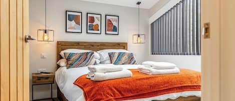 Ein Schlafzimmer mit einem Holzbett, einem Nachttisch, einem Beistelltisch, einem Fenster mit Jalousien und drei gerahmten Kunstwerken an der Wand.