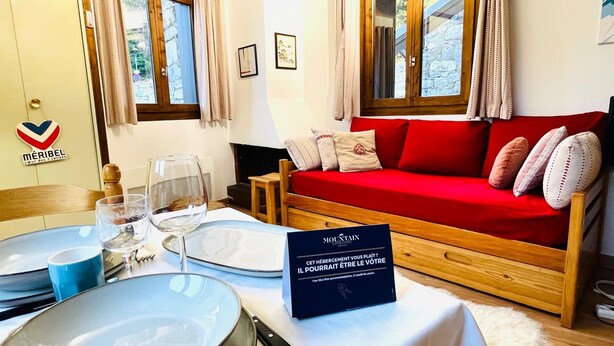 Une pièce chaleureuse avec un canapé rouge, une table à manger dressée avec des assiettes, des verres et des couverts, et une pancarte en français.