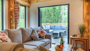 Uma sala de estar acolhedora com um sofá, uma mesa de centro com frutas e uma garrafa, e vista para um deck de madeira e árvores.