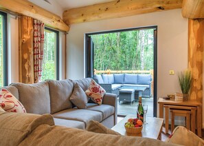 Un salon chaleureux avec un canapé, une table basse sur laquelle se trouvent des fruits et une bouteille, et une vue sur une terrasse en bois et des arbres.