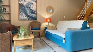 A living room with a blue sofa, wicker chairs, a wooden coffee table, a large wave painting, a round mirror, and a staircase. - Charming 2-Bedroom Cottage Near Guérande and La Baule Beaches, with Terrace and WiFi (Guérande)