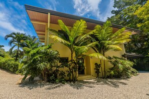 A yellow building with a sloped roof, surrounded by lush greenery and palm trees.