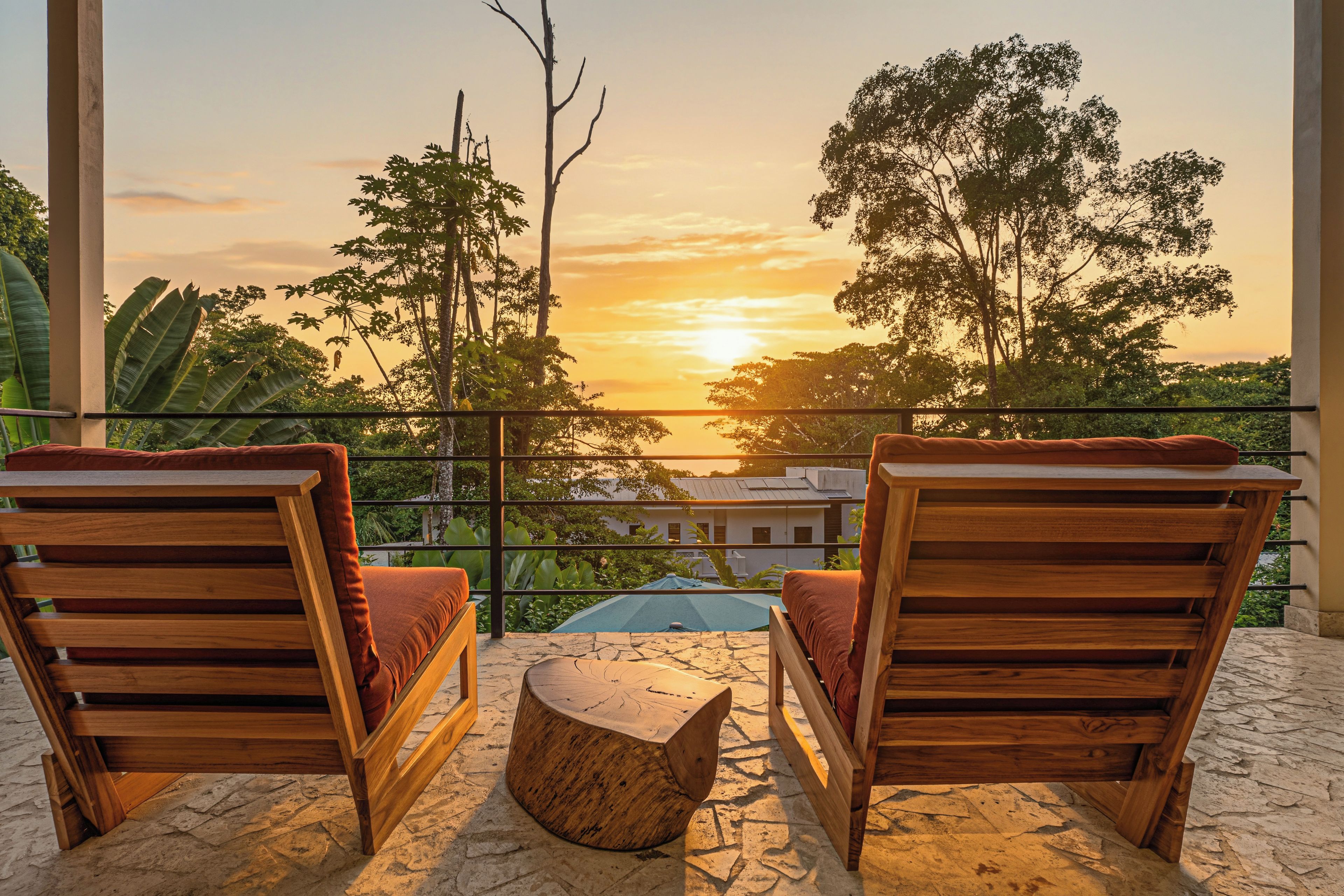 A balcony with two wooden chairs and a small wooden table, overlooking a sunset with trees and buildings in the background.