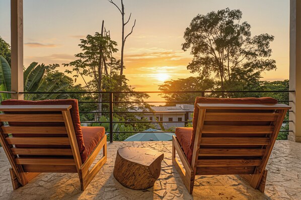 A balcony with two wooden chairs and a small wooden table, overlooking a sunset with trees and buildings in the background.